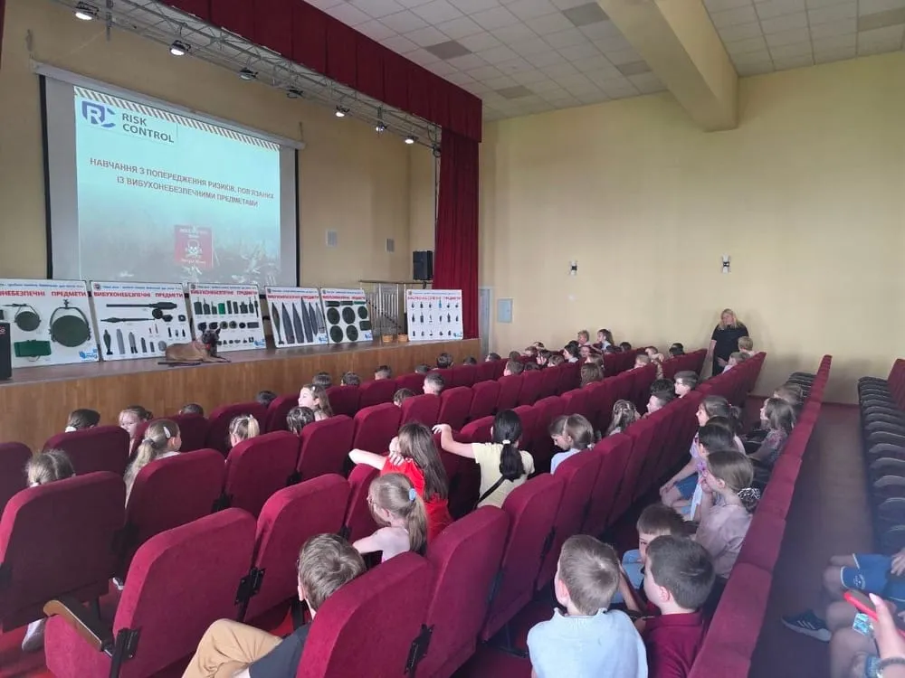 Заняття з мінної безпеки: презентація на екрані та стенди з макетами вибухонебезпечних предметів у залі зі школярами